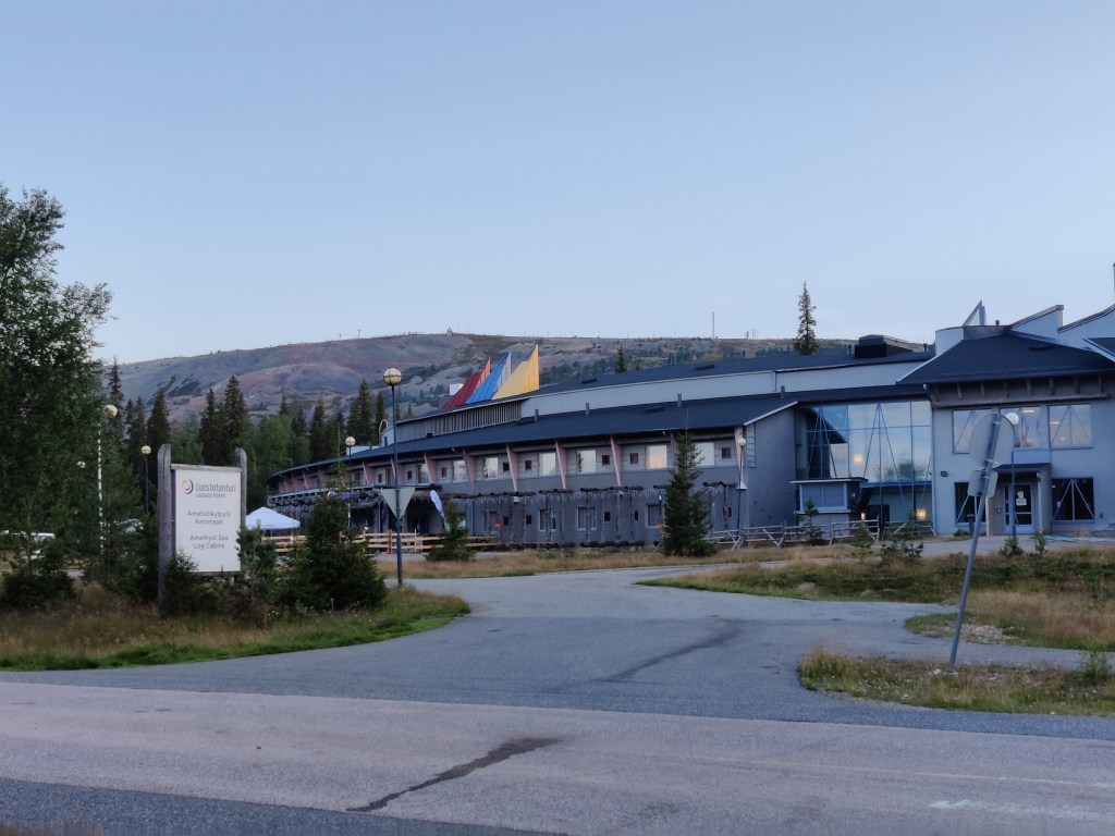 Kuva tienhaarasta, joka vie taustalla seisovalle matalalle hotellille. Hotellin takana näkyy tunturi.