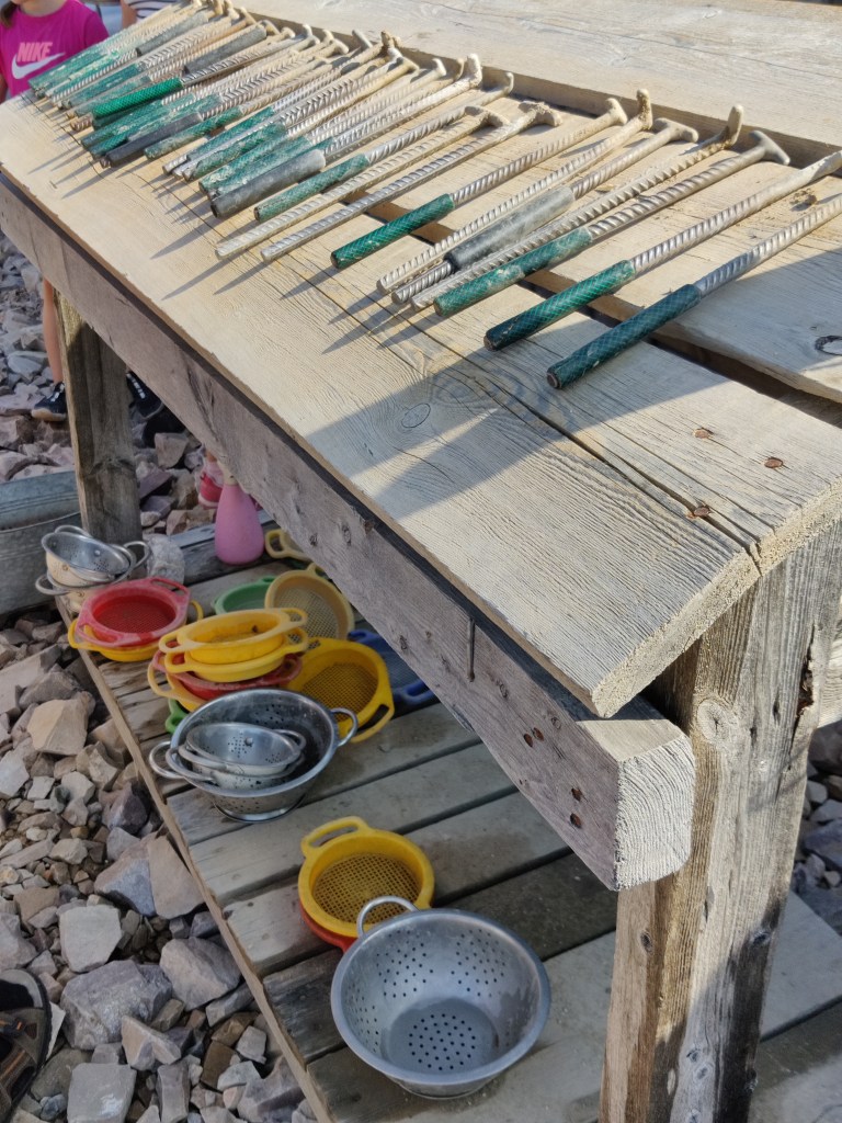 Kuva puisesta telineestä, jolla on rivissä pieniä hakkuja. Telineen alla on erivärisiä siivilöitä.