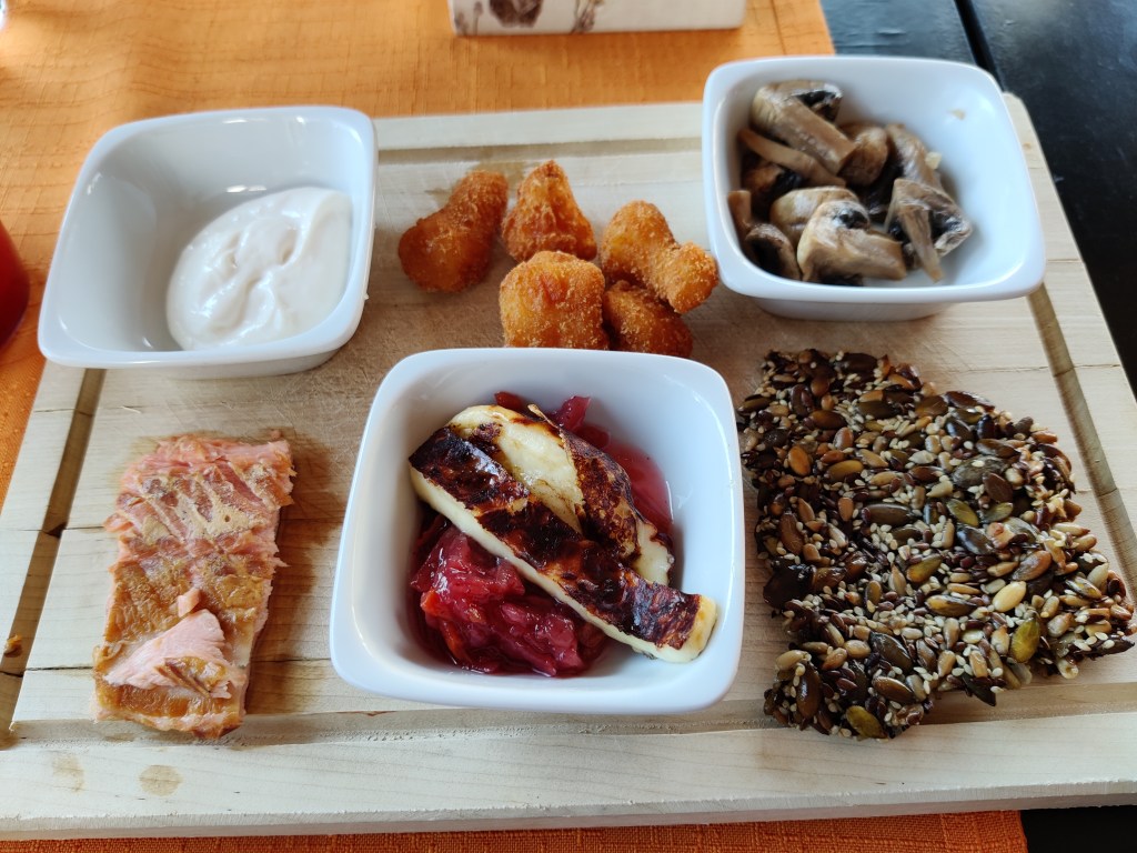 Kuva tarjottimesta, jolla useita pieniä annoksia: majoneesia, lämminsavulohta, kukkakaaliwingsejä, hekkusieniä, siemenleipää ja juustoa sipulihillokkeella.