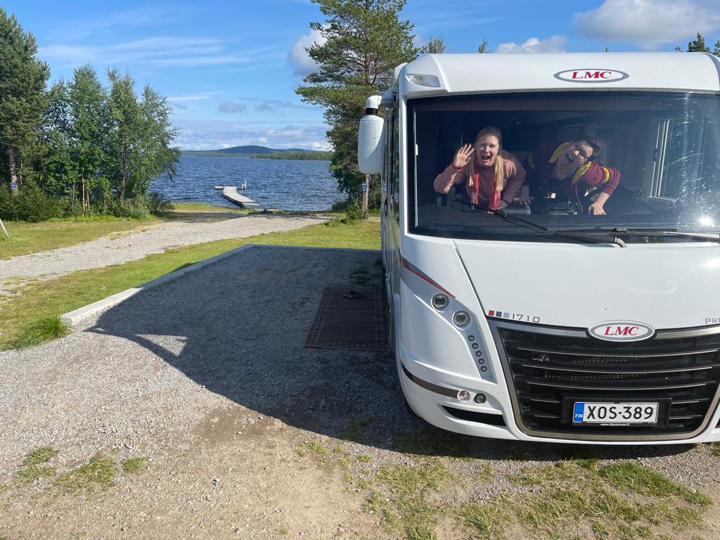 Kuva leirintäalueella seisovan asuntoauton keulasta. Taustalla laituri ja järvi. Auton tuulilasin läpi näkyy kaksi ilveilevää ihmistä. 
