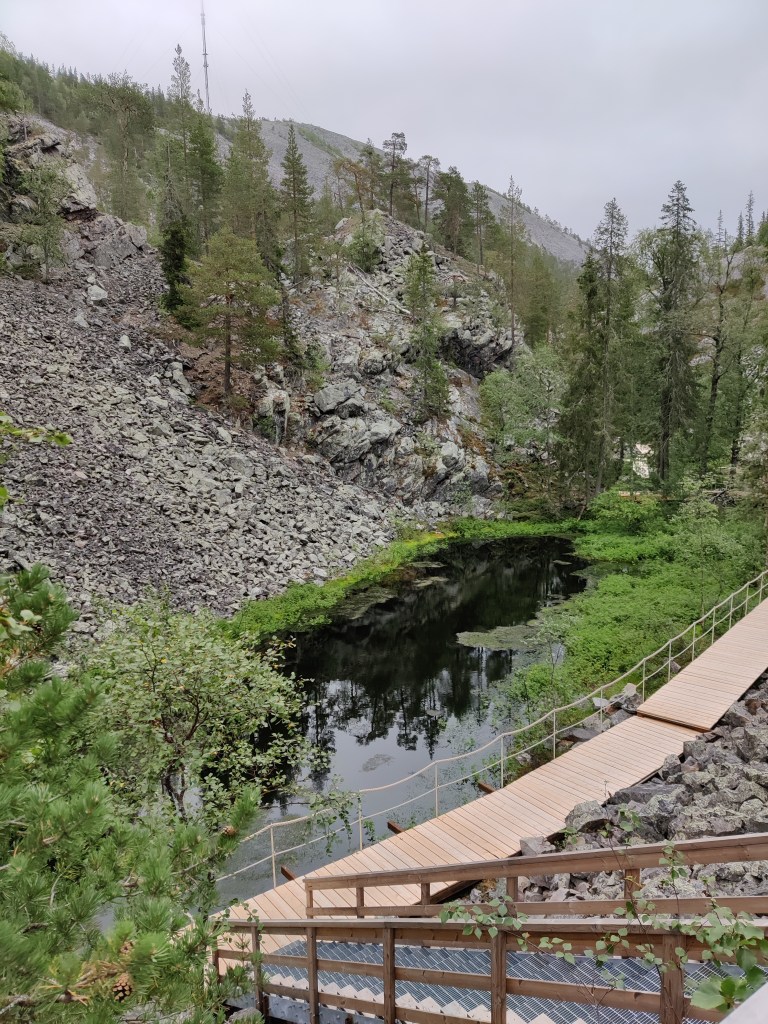 Kuva rappusilta alaspäin kävelytasanteelle, jonka vieressä on pieni lampi kivikkoisen kallion juurella. Havupuita taustalla.