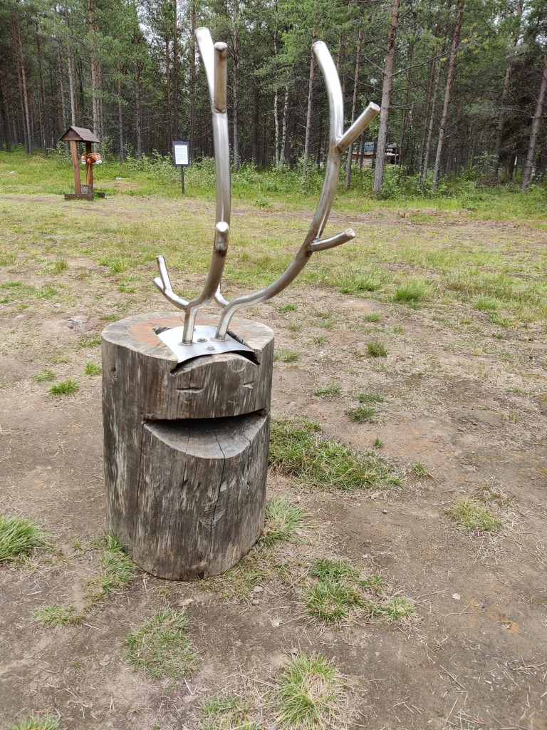 Kuva puupöllistä, jonka päälle on pultattu metalliset poronsarvet. Pölliin on kaiverrettu silmät ja avoin suu.