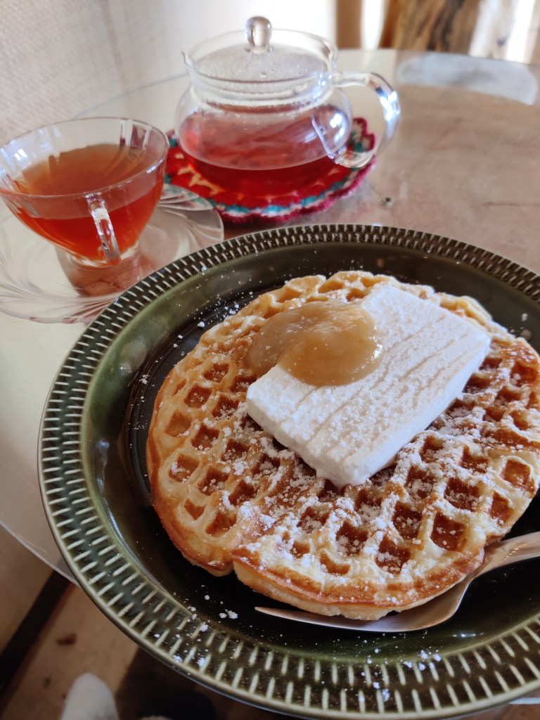 Erikoislähikuva lasisesta teepannusta ja teekupista lautasineen sekä lautasellisesta vohvelia, jonka päällä on vaniljajäätelöä ja omenahilloa.