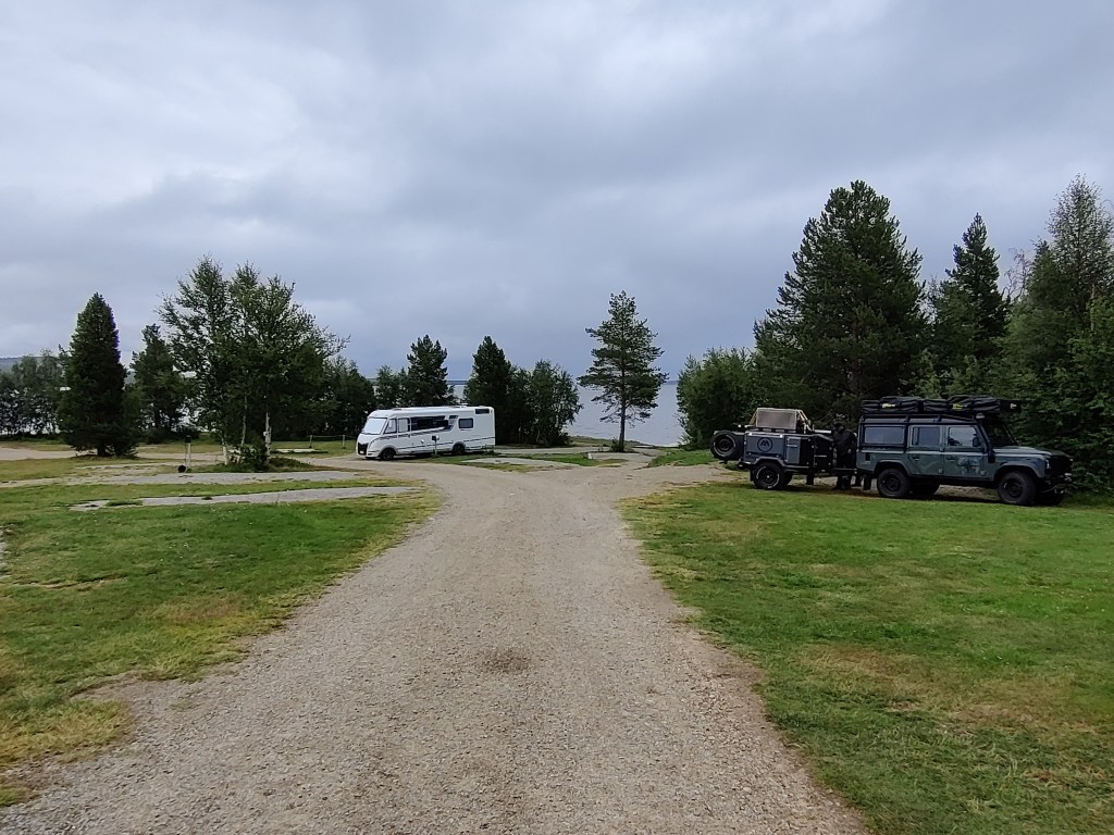 Iso kuva leirintäalueesta: hiekkatie vie suoraan eteenpäin. Kuvassa näkyy valkoinen, bussimainen asuntoauto sekä tumma maasturi, jossa on omituinen peräkärry. Taustalla järvimaisema.