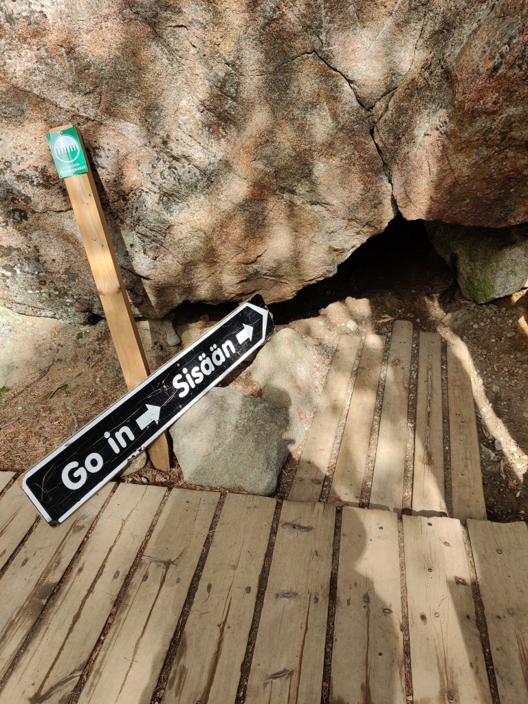 Kuva, jonka etualalla on puisista lankuista tehty seisoma-alue ja kyltti, jossa lukee "Go in (nuoli) Sisään (nuoli)". Kyltti osoittaa ison kiven alla olevaan pieneen aukkoon.