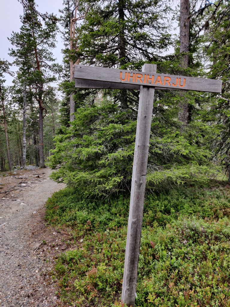 Lähikuva puisesta kyltistä, jossa lukee "Uhriharju". Taustalla varpuja, havupuita ja hiekkapolku.