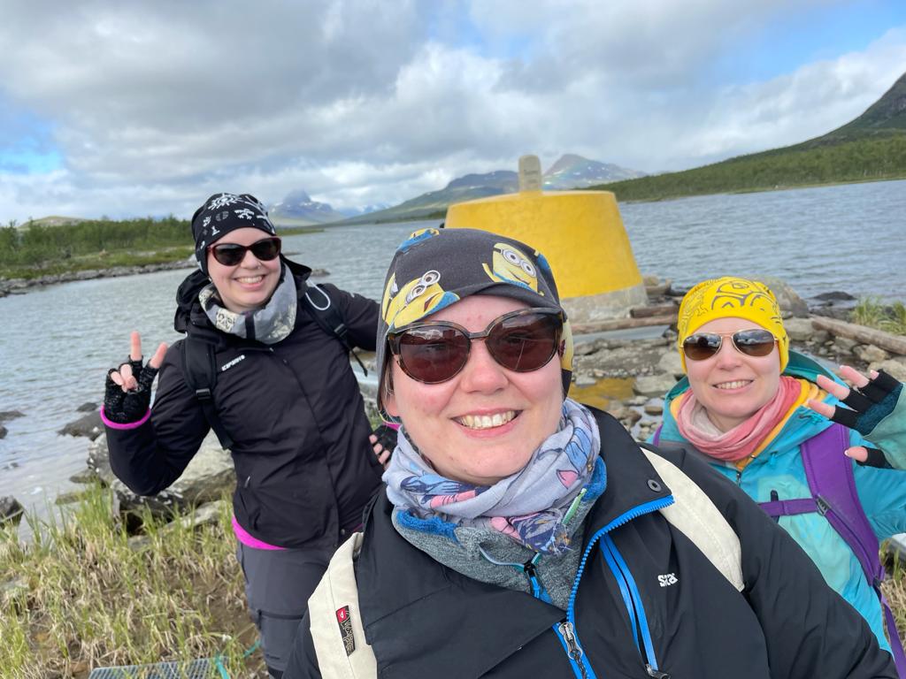 Selfie Katjasta, Tuulasta ja Tainasta Kolmen valtakunnan rajapyykin edessä. Kaikilla pipot, aurinkolasit ja onnellinen hymy.