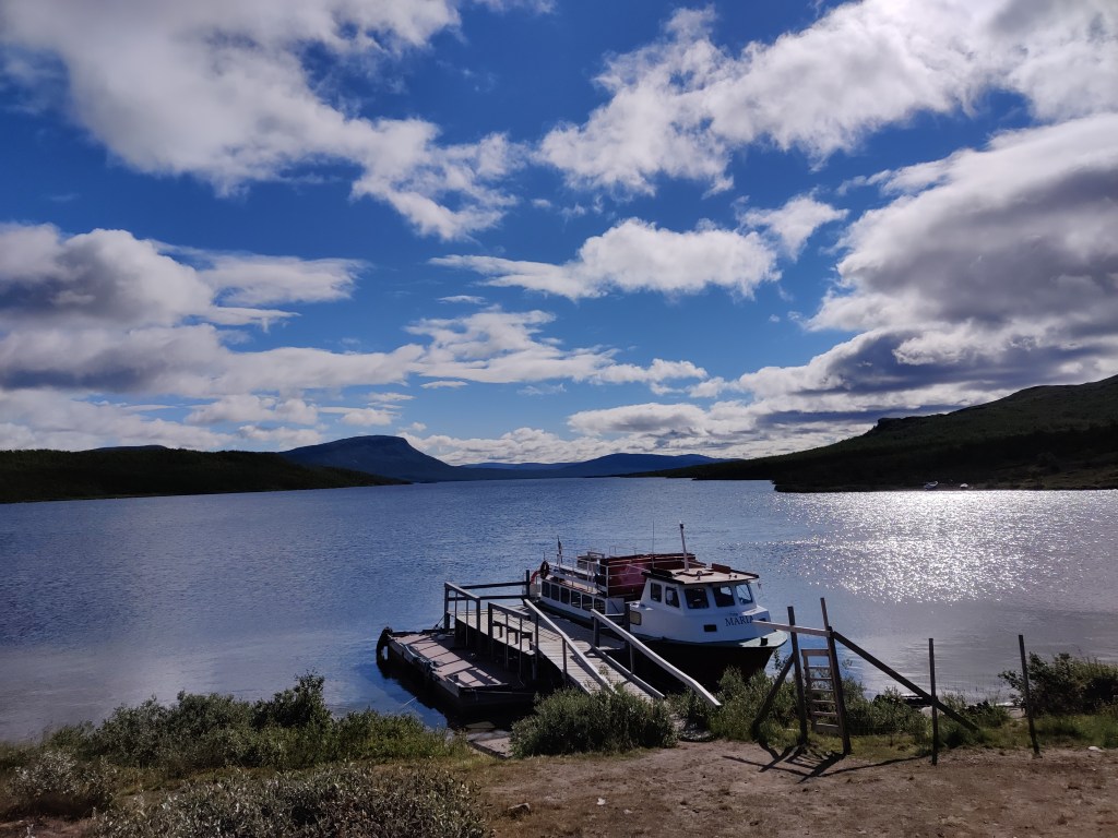 Kuva järvimaisemasta, jonka taustalla siirtää tuntureita. Rannassa on puinen laituri ja iso koppivene.