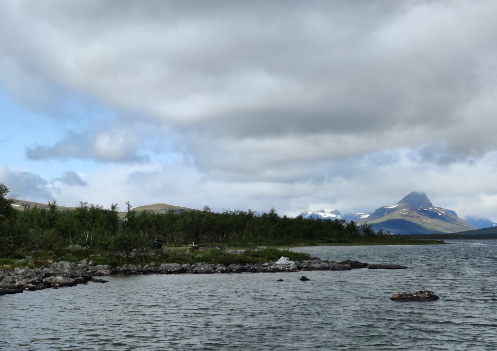 Järvimaisema, kivikkoinen ranta ja kitukasvuisia koivuja. Kuvan oikeassa reunassa hain evää muistuttava vuori kohoaa pilviin, jotka roikkuvat matatalla.