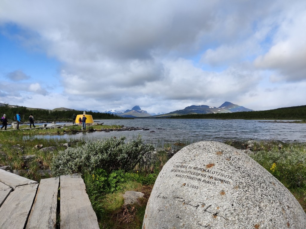 Kuva Kolmen valtakunnan rajapyykistä, joka erottuu keltaisena rakennelmana järven reunalta. Sinne vie puiset pitkospuut. Etualalla muistokivi, johon on kirjoitettu kolmella kielellä Kalottireitin avaus ja päivämäärä. Kuvan taustalla tunturimaisema.