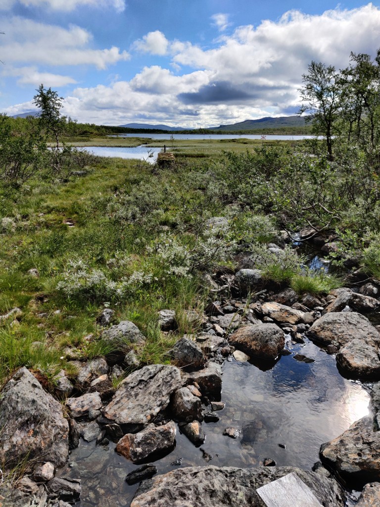 Kuva karusta maisemasta, jossa kiviä, vettä ja varpuja. Keskellä kuvaa näkyy keltaiseksi värjätty merkki, joka erottaa Suomen ja Ruotsin valtioiden rajat toisistaan.