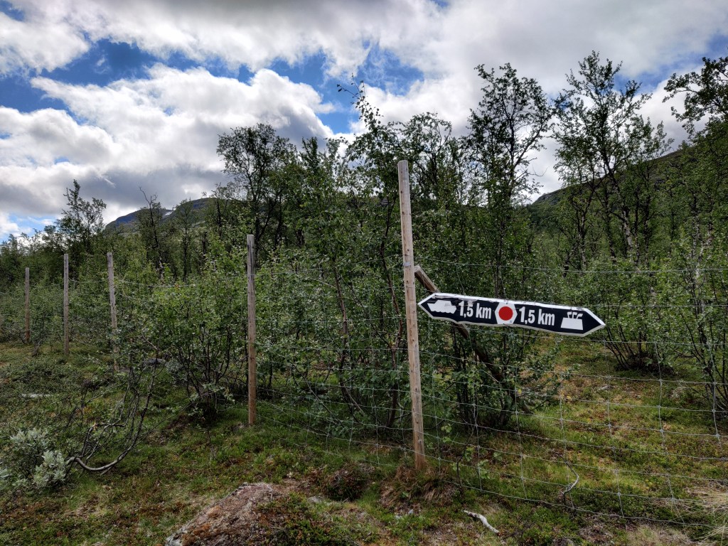 Kuva maaston keskellä olevasta verkkoaidasta, jossa on selkeä kyltti. Kyltin vasen puoli osoittaa kohti rantaa eli siinä on vene ja teksti 1,5 km. Toinen puoli osoittaa rajapyykille eli oikealle ja siinä lukee myös 1,5 km.