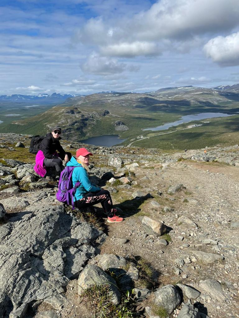 Kuva Katjasta ja Tainasta istumassa kivillä polun vieressä. Rinne on jo melko korkea ja taustalla olevaa järveä voi jo katsoa yläviistosta. Aurinko paistaa, mutta taivaalla on pilviä.