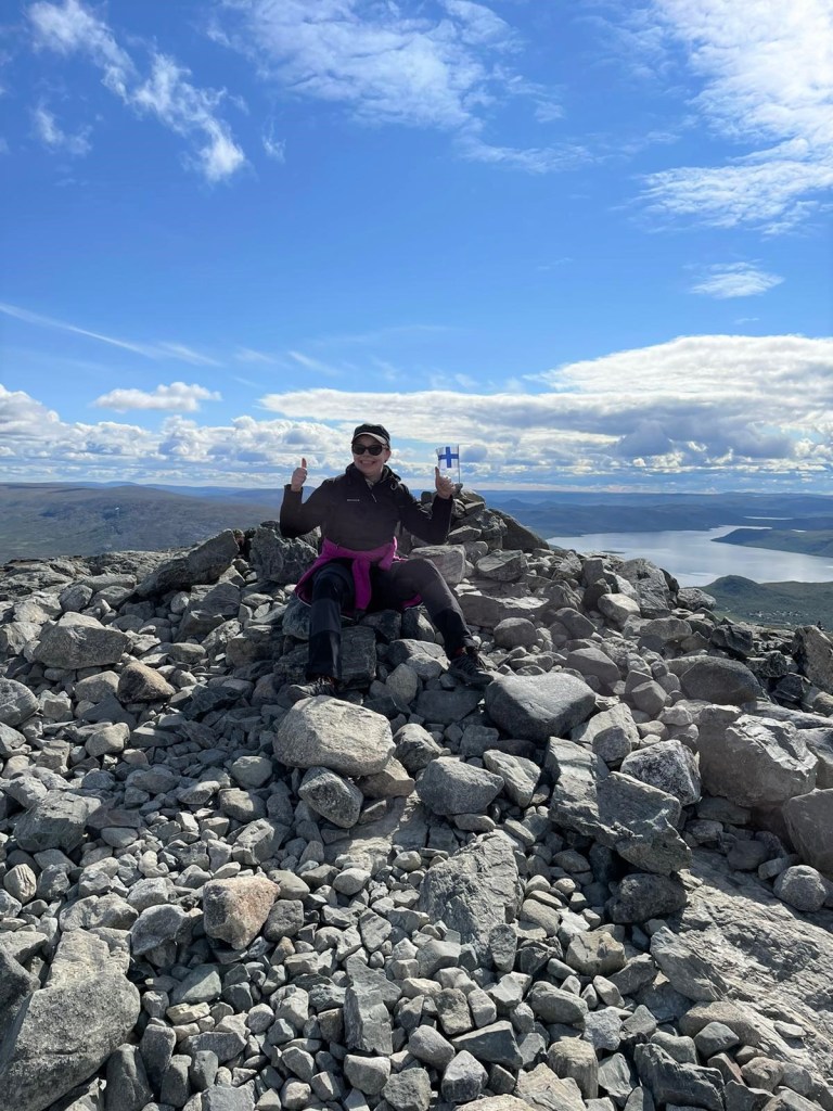 Kuva Katjasta näyttämässä peukkuja istuskellessaan Saanan kivikkoisella huipulla. Takana liehuu pieni SUomen lippu. Taustalla järvii ja muita tuntureita. Aurinko paistaa.