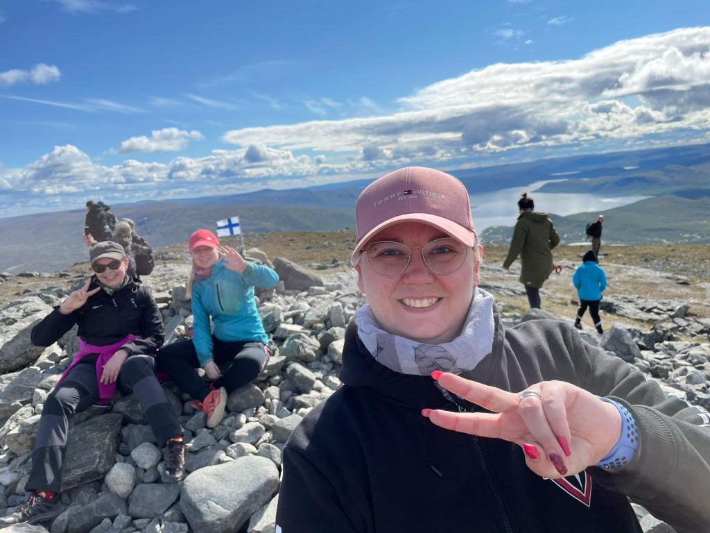 Selfie Katjasta, Tainasta ja Tupusta, jotka moikkaavat kameralle Saanan huipulla. Huippu on kivikkoinen ja sen päällä on pieni Suomen lippu. Taustalla muita ihmisiä.