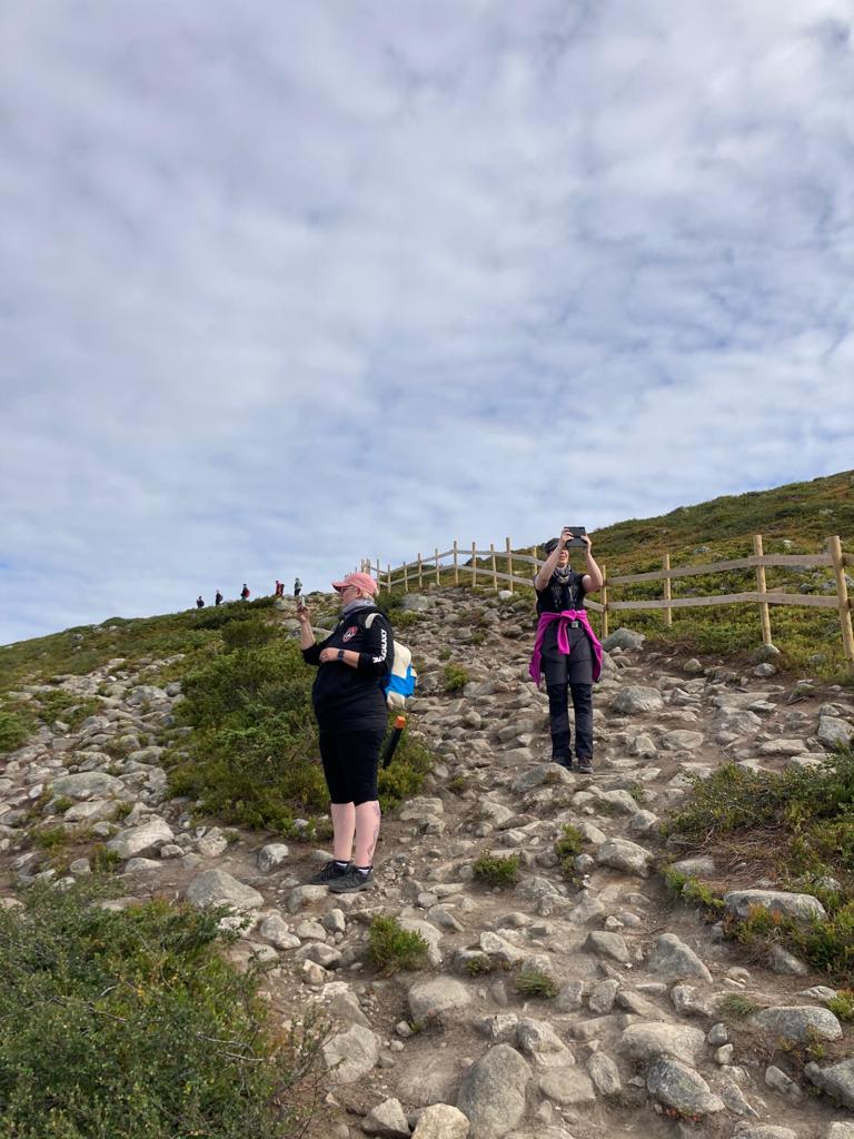 Kuva Katjasta ja Tuulasta rinteessä räpsimässä kuvia puhelimilla. Rinteessä kivikkoinen polku ja varpuja. Taka-alalla puinen aita ja kaukana rinteessä ihmisiä.