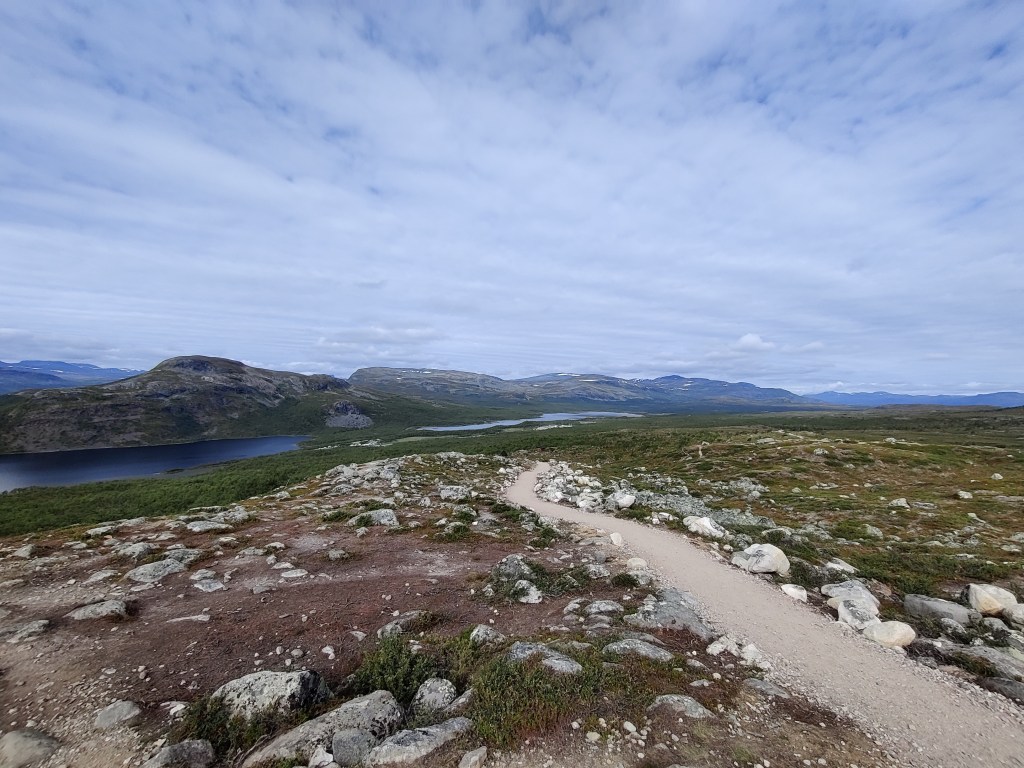 Maisemakuva, jossa etualalla kivikkoinen maasto ja hiekkapolku. Taustalla näkyy kapea järvi ja vastakkaisella rannalla tuntureita.