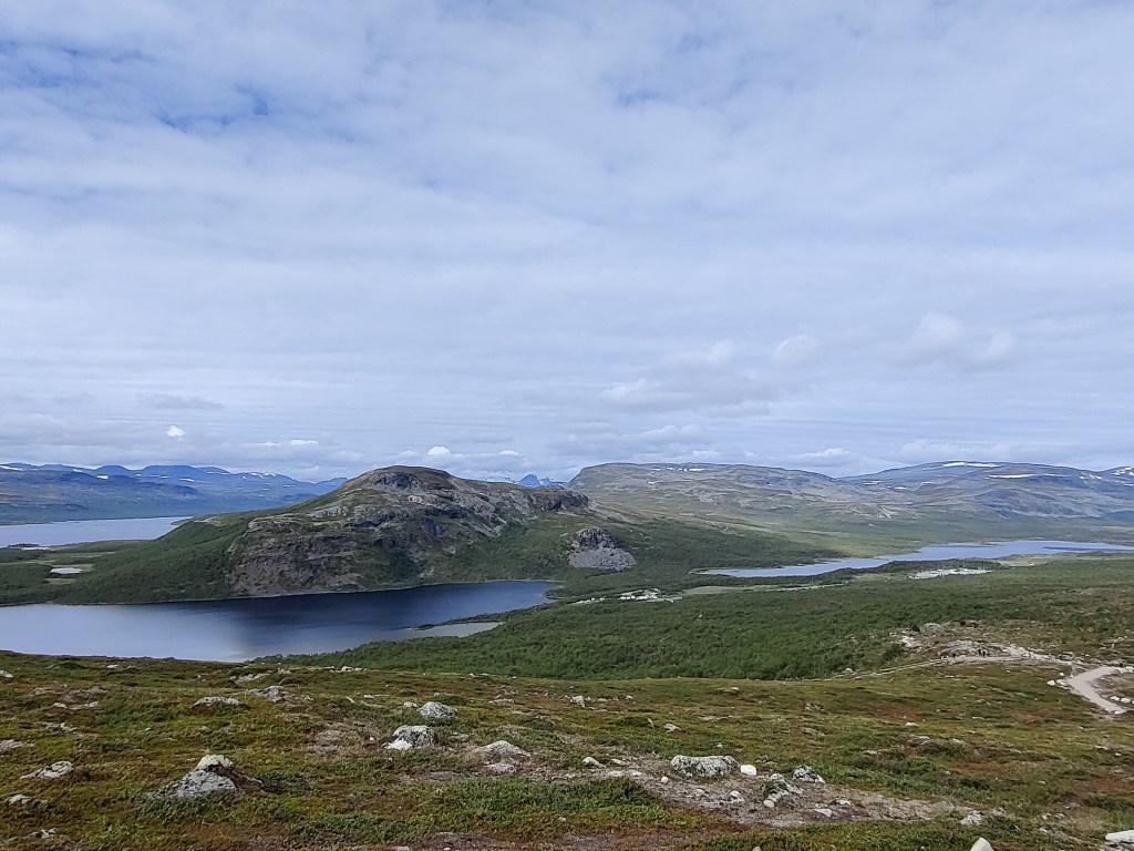 Maisemakuva hieman ylempää rinteestä: maasto on sammalista ja varpuista. Järvi erottuu selkeämmin ja joillain tuntureilla rannan toisella puolella on jopa hieman lunta.