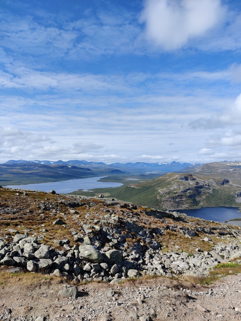 Maisemakuva, jossa etualalla hyvin kivikkoinen rinne. Taustalla näkyy järvi, tuntureita ja lisää kaukana siintäviä tunteita. Taivas on osittain pilvinen.