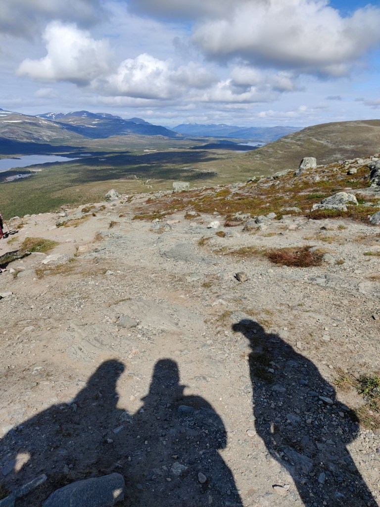 Maisemakuva rinteeltä alas kohti polkua, jota pitkin on noustu. Kivikkoa ja karua maastoa. Kolmen ihmisen varjot heijastuvat polulle.