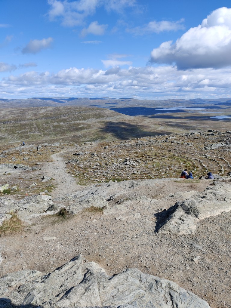 Maisemakuva Saanan huipulta. Huipun vieressä on ihmisen tekemä kivistä kasattu kehä, jonka viertä kulkee hiekkainen polku. Taustalla näkyy tunturimaisema. Pilvien varjot heijastuvat maastoon. Satunnaiset ihmiset istuvat rinteessä tauolla.