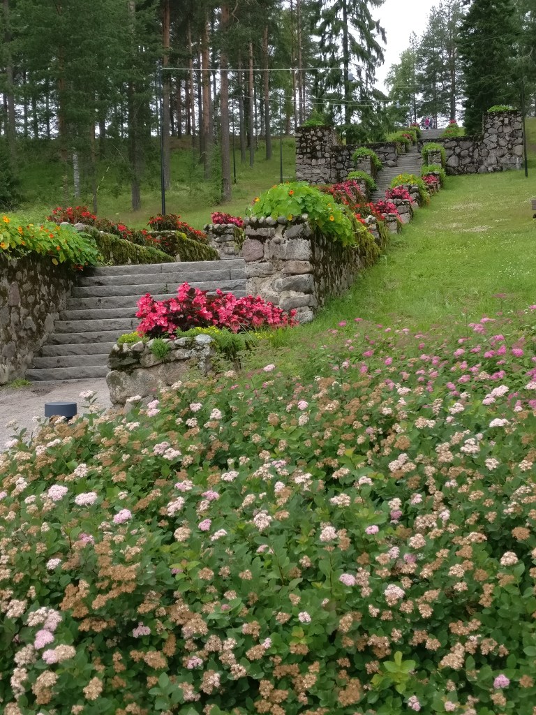 Kuva kivisistä portaista, jotka kohoavat puisen harjun juurelle. Portaita reunustavat kivikaiteet ja runsas määrä punaisia ja oransseja kukkia. Nurmikko vihertää ja etualalla on iso kukkapensas.