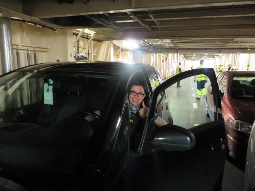 Kuva Katjasta sisällä autolautassa: hän istuu autossa, ovi on auki ja näyttää kuvaajalle peukkua. Kasvoilla on leveä hymy.