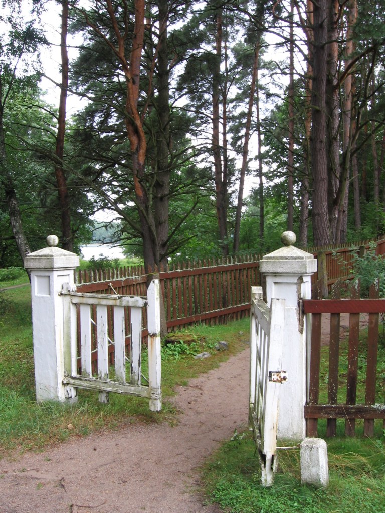 Kuva hautausmaan portista. Portti mataline pylväineen on maalattu valkoiseksi, muu osa puista aitaa on punertavan ruskea. Portti on auki ja sinne menee hiekkainen polku. Taustalla metsikköä, jonka läpi siintää meri.