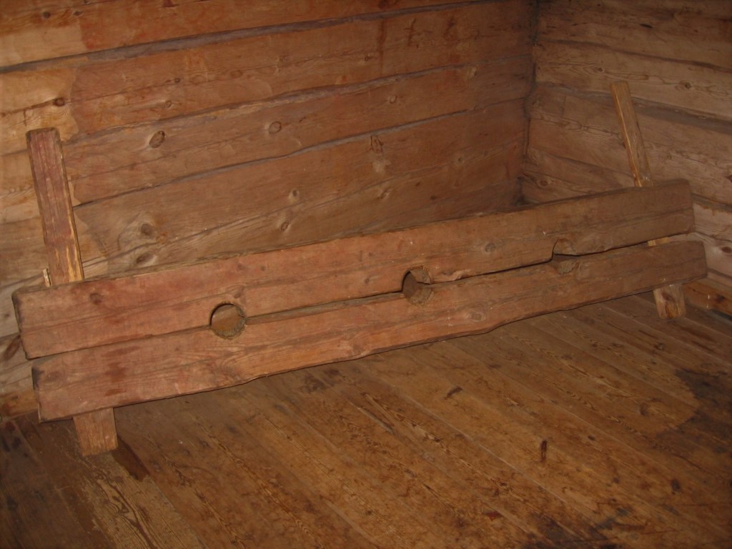 Kuvassa puukirkon nurkkaus, jossa esillä puinen jalkapuu, jossa kolme reikää: yksi päälle ja kaksi käsille.