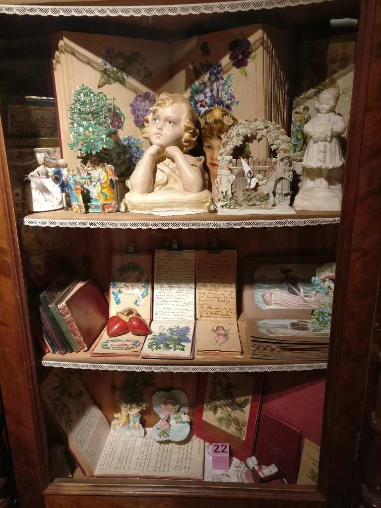 Kuva vitriinikaapista, jossa koriste-esineitä ja kiiltokuvin koristeltuja runovihkoja.