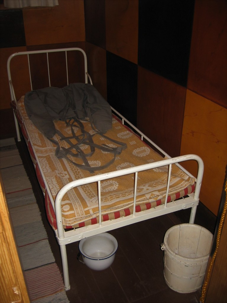 Erikoislähikuva valkoisesta terässängystä, jolla lojuu vanha pakkopaita. Sängyn etupuolella on yöastia ja valkoiseksi maalattu puinen sanko.