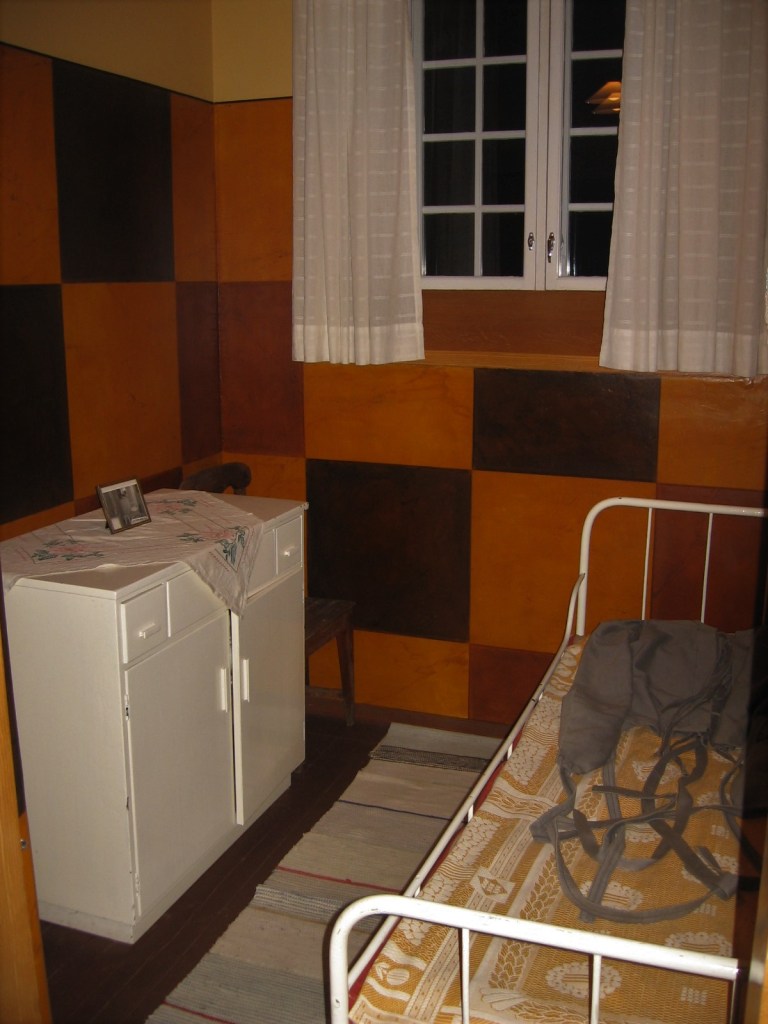 Kuva ahtaasta huoneesta, jossa on valkoinen matala kaappi, räsymatto ja valkoinen terässänky. Sängyllä on tummanharmaa pakkopaita. Huoneen seinissä on erivärisiä neliöitä. Valkopuitteisessa ikkunassa on valkoiset ikkunaverhot.