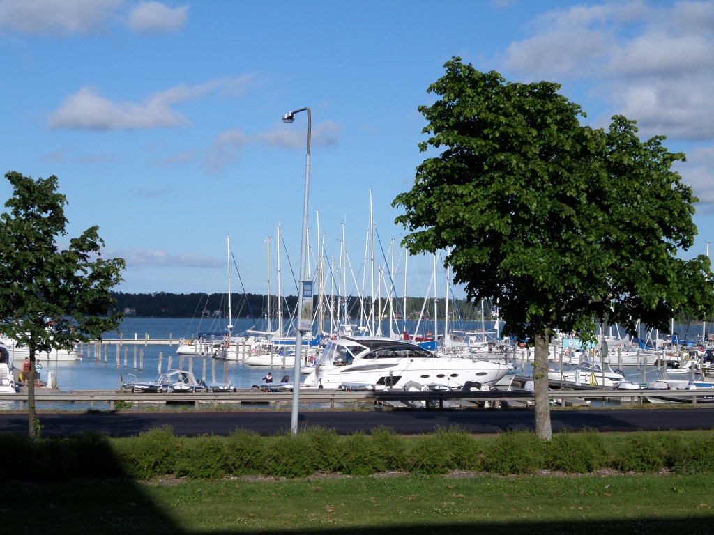 Kuva satamasta: etualalla nurmikenttä ja kaksi lehtipuuta. Satamassa lukuisia valkoisia veneitä, joista valtaosa purjeveneitä. Edessä yksi pikavene.