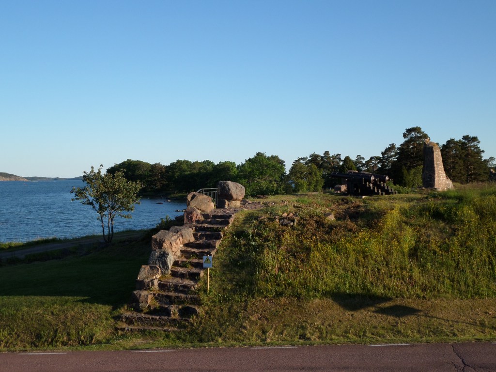 Kuva rannasta, jossa näkyy kukkulalla linnoituksen kivisiä portaita ja kauempana lisää vanhoja muureja sekä tykki.