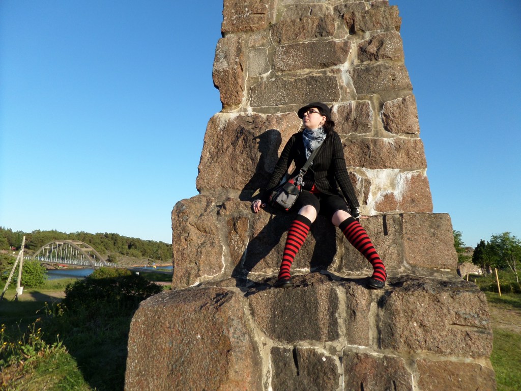 Kuva korkeasta kivimuurista, jonne Katja on kiivennyt istumaan. Muurin huippu ei mahdu kuvaan. Taustalla vilkkuu meri ja kaarisilta.