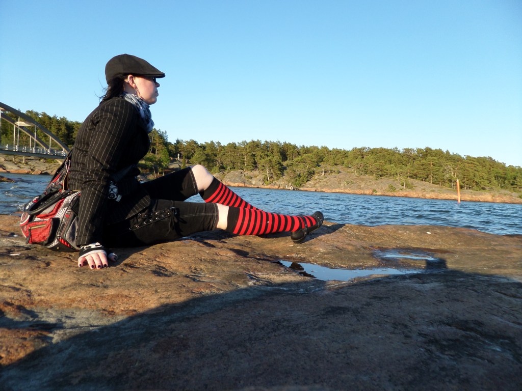 Kuva Katjasta, joka istuu matalalla rantakalliolla ja tuijottaa merta. Punamustaraidalliset sukat erottuvat muusta mustasta asukokonaisuudesta.