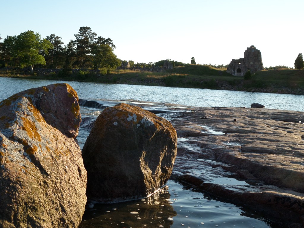 Kuva merenrannalta: etualalla kiviä, taustalla näkuu ranta, jossa linnoituksen raunioita. Takavasemmalla lehtipuita.