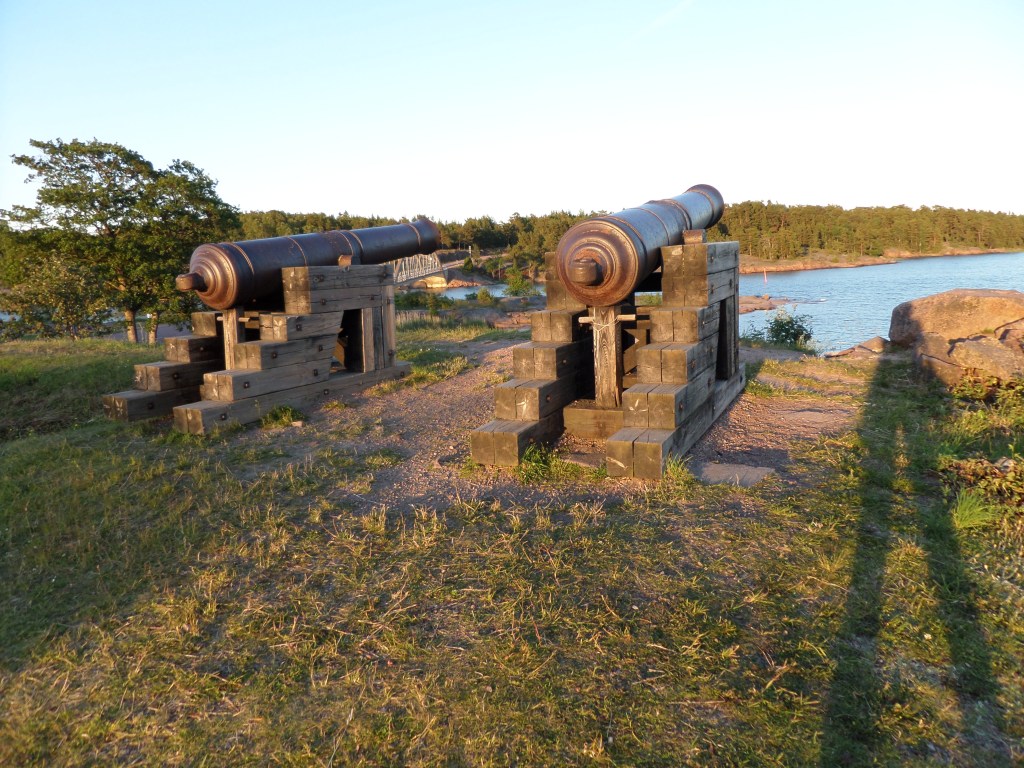 Kuva kahdesta tykistä, jotka osoittavat meren suuntaan. Aurinko paistaa kuvaajan takaa ja tämän varjo heijastuu tykkien viereen.