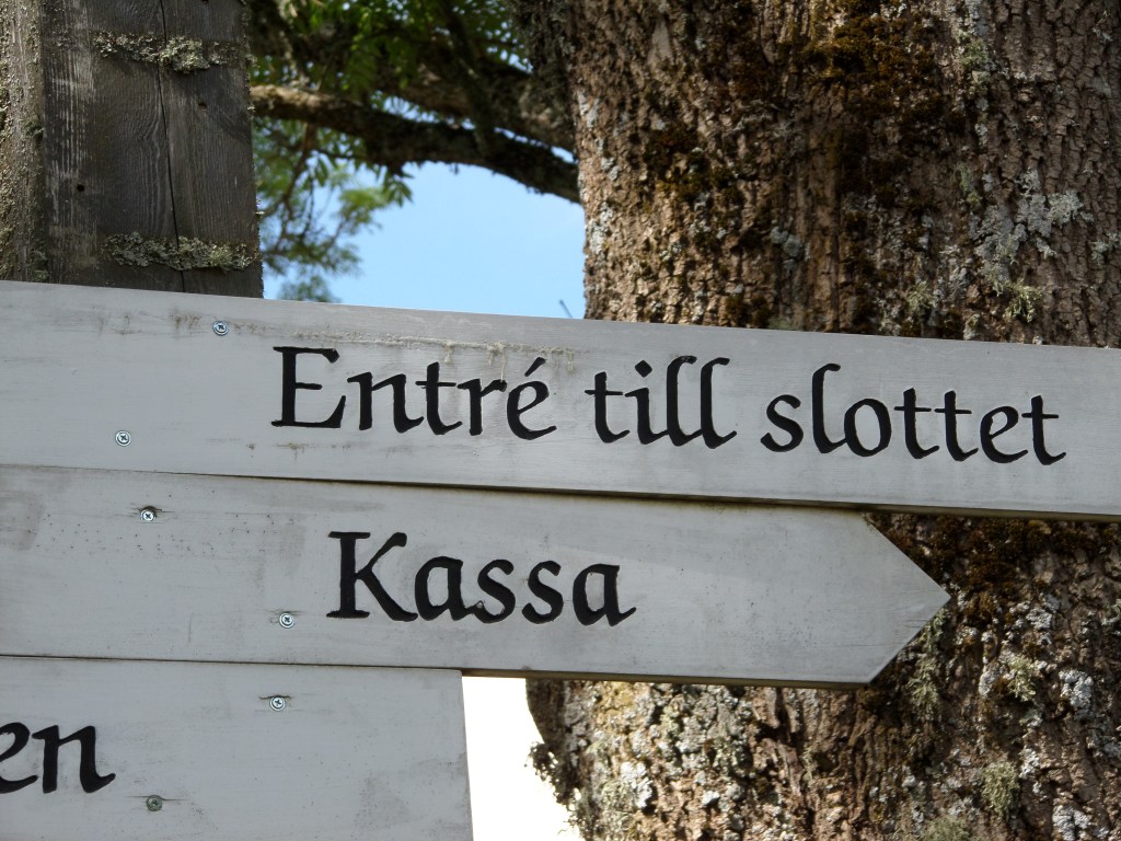Erityislähikuva puisesta kyltistä, jossa lukee "Entré till slottet" ja "Kassa".