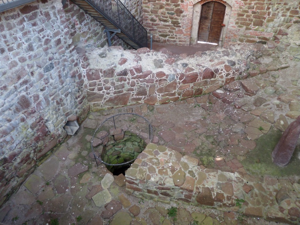 Kivinen sisäpiha, jolle pääsee laskeutumaan moderneja rappuja pitkin. Kivisessä maassa on pyöreä kaivo, joka on aidattu raitaisella kehikolla.