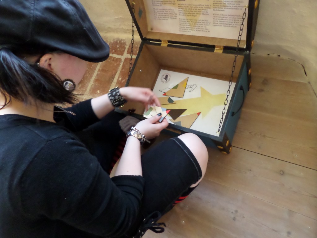 Niskan takaa otettu kuva Katjasta, joka kasaa puisessa laatikossa ollutta pulmapalapeliä, josta pitäisi muodostaa mallin mukainen kuvio.