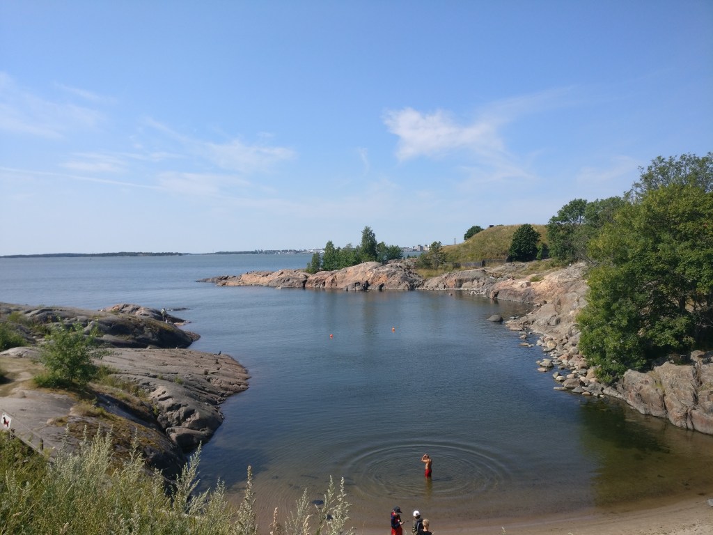 Kuva alhaalla olevasta pienestä lahdesta, jossa on hiekkapohjainen uimaranta. Rantaa reunustavat kiviset kalliot. Muutamia ihmisiä näkyy rannalla.