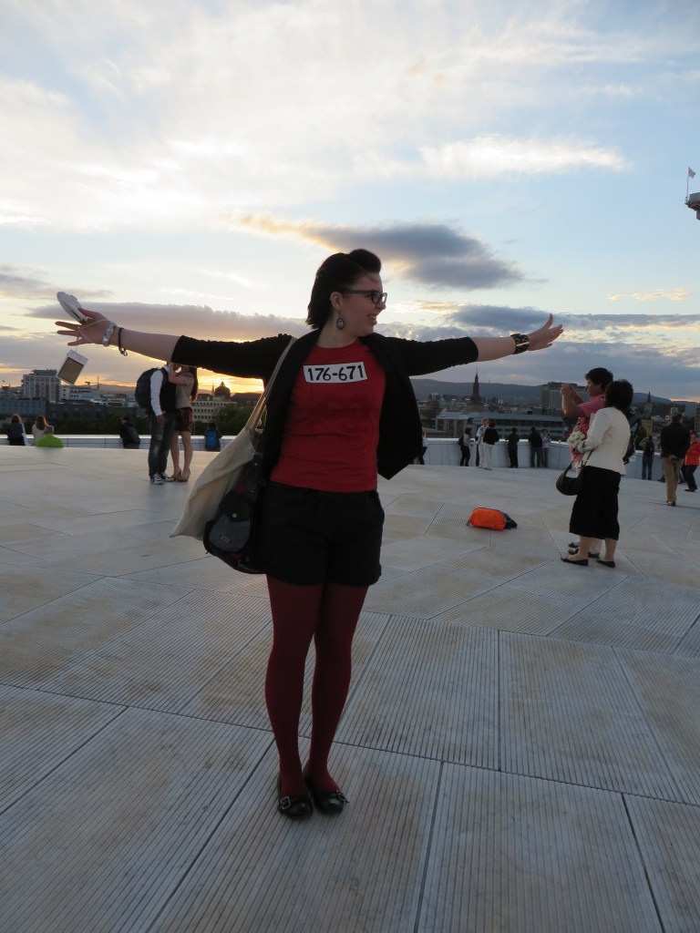Kuva Katjasta oopperatalon kattotasanteella levittämässä käsiään kuin olisi lähdössä lentoon. Katjalla on mustat shortsit, punaiset sukkahousut ja punainen Karhukopla-t-paita, jossa lukee 176-671.
Taustalla useita turisteja.