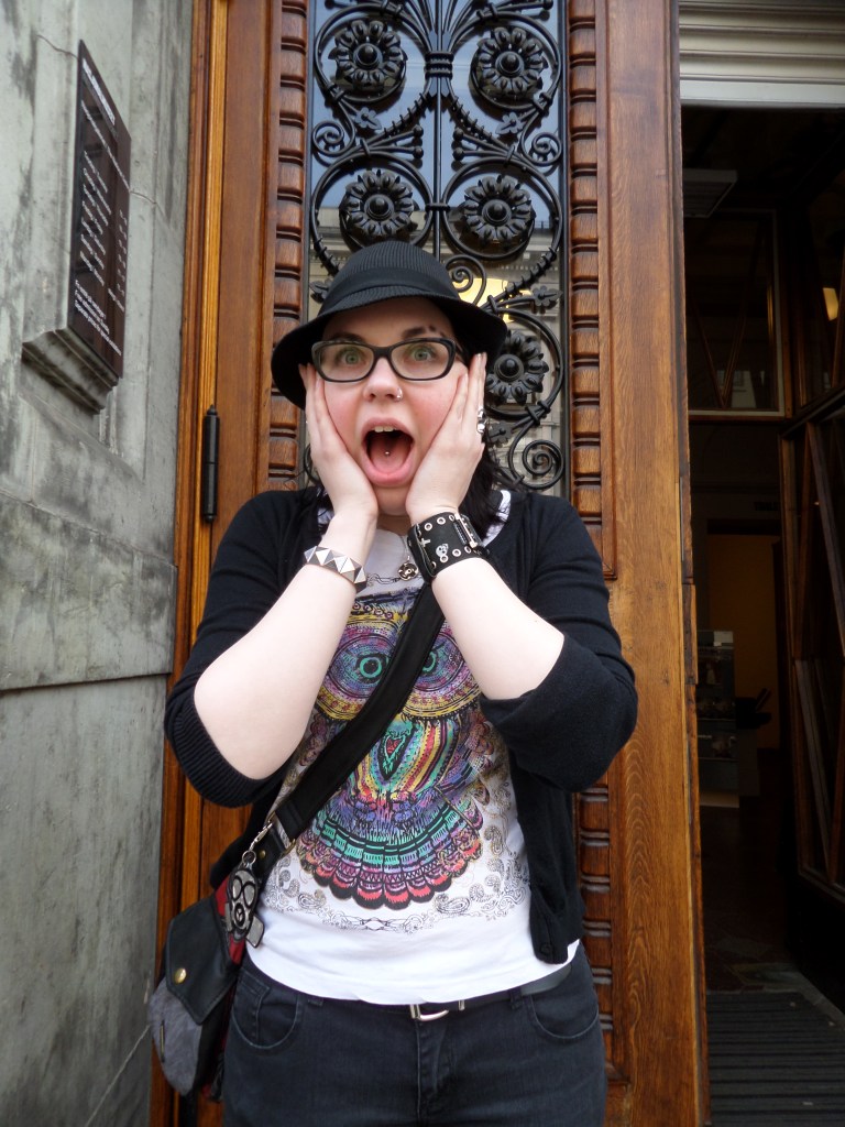 Kuva Katjasta, joka poseeraa museon oven vieressä kädet poskillaan, suu auki, leikkien Munchin Huuto-maalausta. Katjalla on musta hattu, musta neulepusero ja valkoinen pöllöpaita päällä.