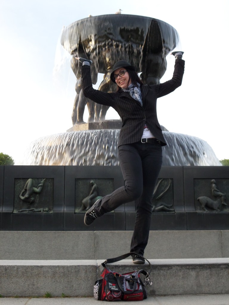 Kokokuva Katjasta poseeraamassa ison suihkulähteen edessä. Suihkulähteen jalustassa on kuvattu ihmishahmoja eri asennoissa ja suihkulähteen maljaa kannattelee myös ihmishahmoja. Katjalla on kädet ja toinen jalka ilmassa. Hänellä on mustat farkut ja musta liituraitatakki sekä musta hattu.