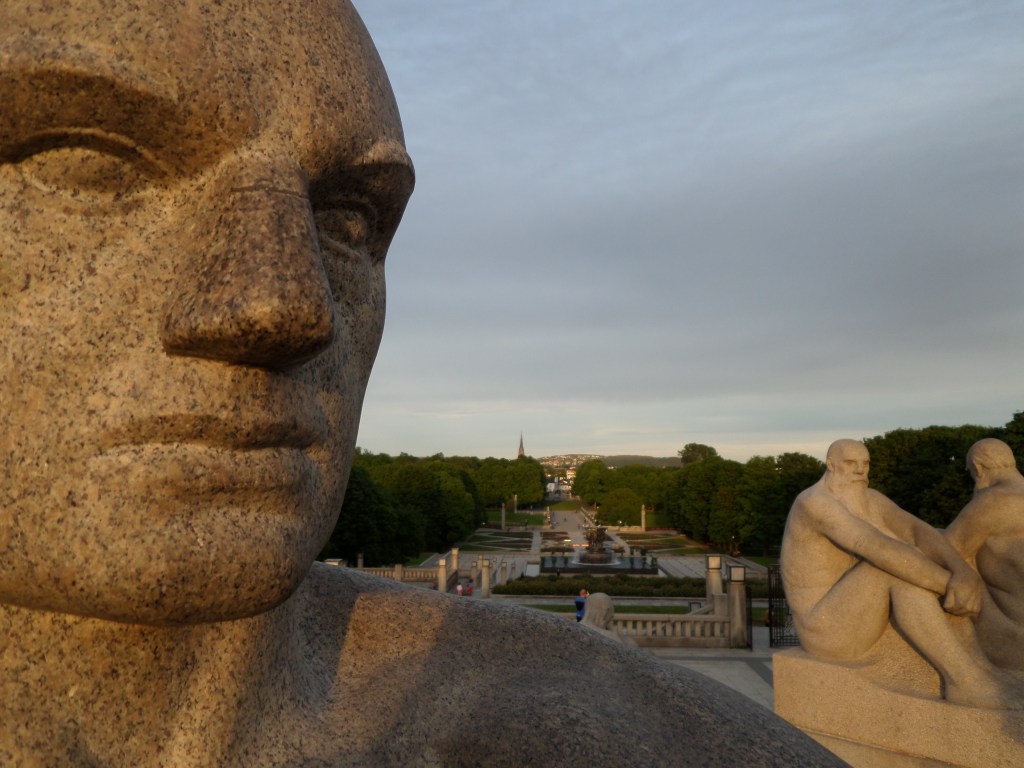 Erityislähikuva ihmispatsaan kasvoista. Taustalla näkyy kaksi muuta patsasta ja keskellä kuvaa näkyy puiston vihreiden puiden reunustaman polun levittäytyminen kauas horisonttiin.