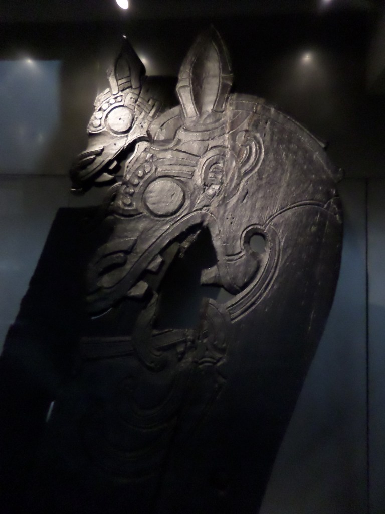 Lasin läpi otettu kuva kahdesta puusta veistetystä lohikäärmeenpäästä, jotka ovat todennäköisesti olleet viikinkilaivojen koristeita.