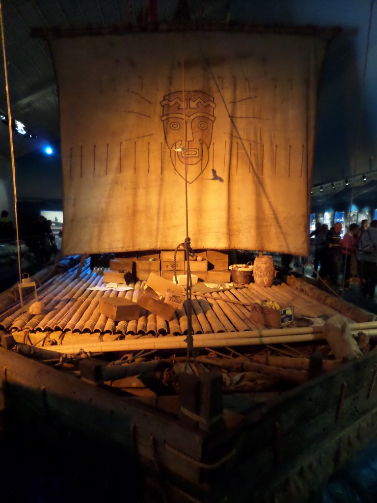 Kuvassa puinen laiva, jonka kanesi näyttää koostuvan jonkinlaisista bambun tai vastaavan rungoista. Kannella on laatikoita ja paljon sekalaista tavaraa. Iso, neliönmallinen purje on avoinna. Siihen on kuvattu ihmispää, jolla on parta. Valaistus on keltainen.