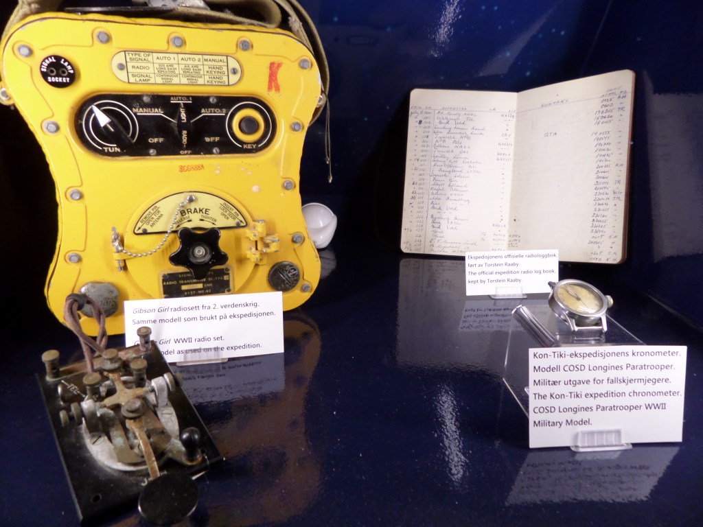 Kuva vitriinissä olevista esineistä: vanha radiosetti, muistikirja ja kelloa muistuttava kronometri.