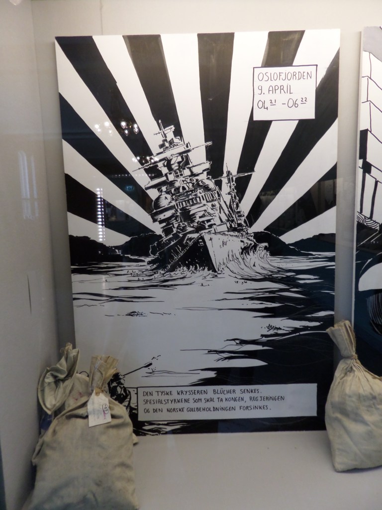 Kuva seinällä olevasta mustavalkoisesta sarjakuvaruudusta, jossa sotalaiva näyttää hitaasti uppoavan veteen. Etualalla on kaksi kangaspussia.